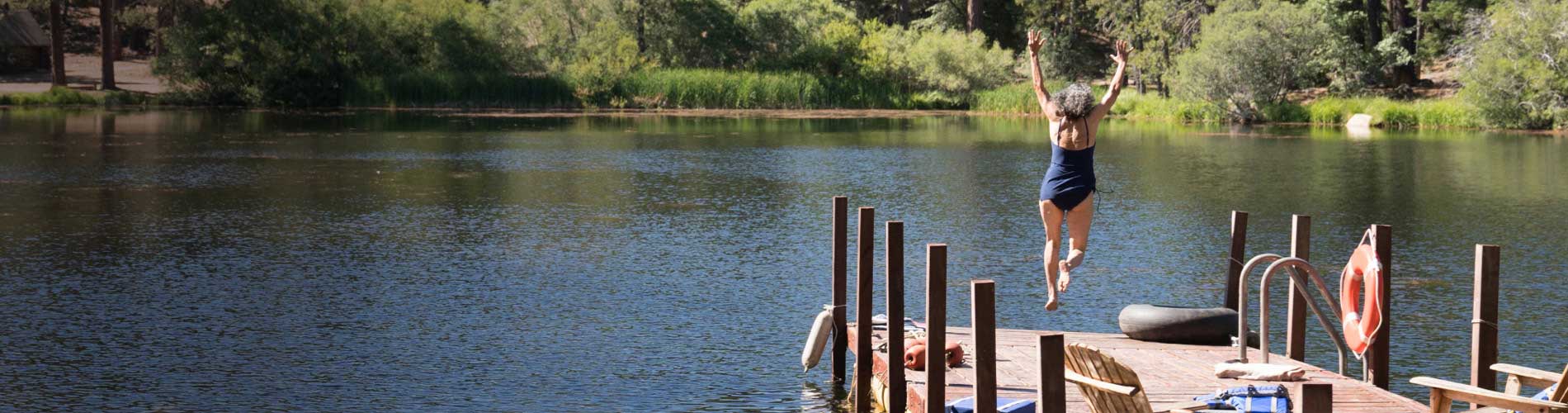 People swimming in a lake surrounded by trees with a dog on the dock.