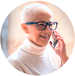Woman with short white hair and blue glasses, wearing a turtleneck is speaking on the phone and smiling.