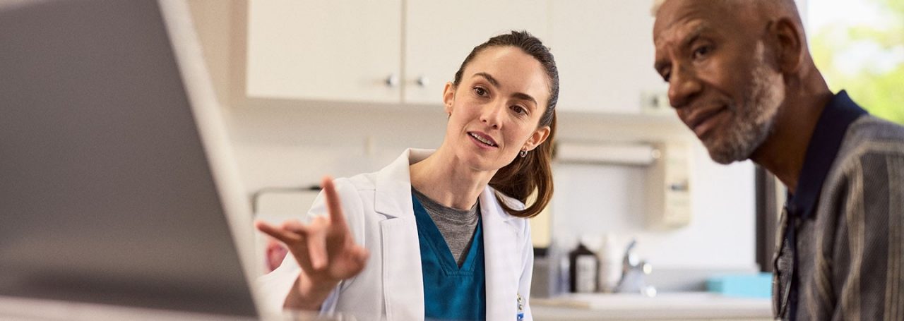 A healthcare professional in white coat pointing to a screen with a patient next to them in an exam room.
