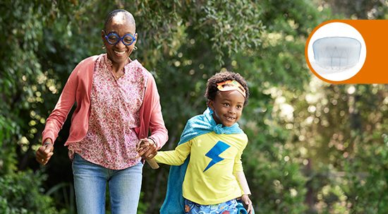 A smiling woman in a pink top and jeans holding hands with a young girl in a superhero costume.