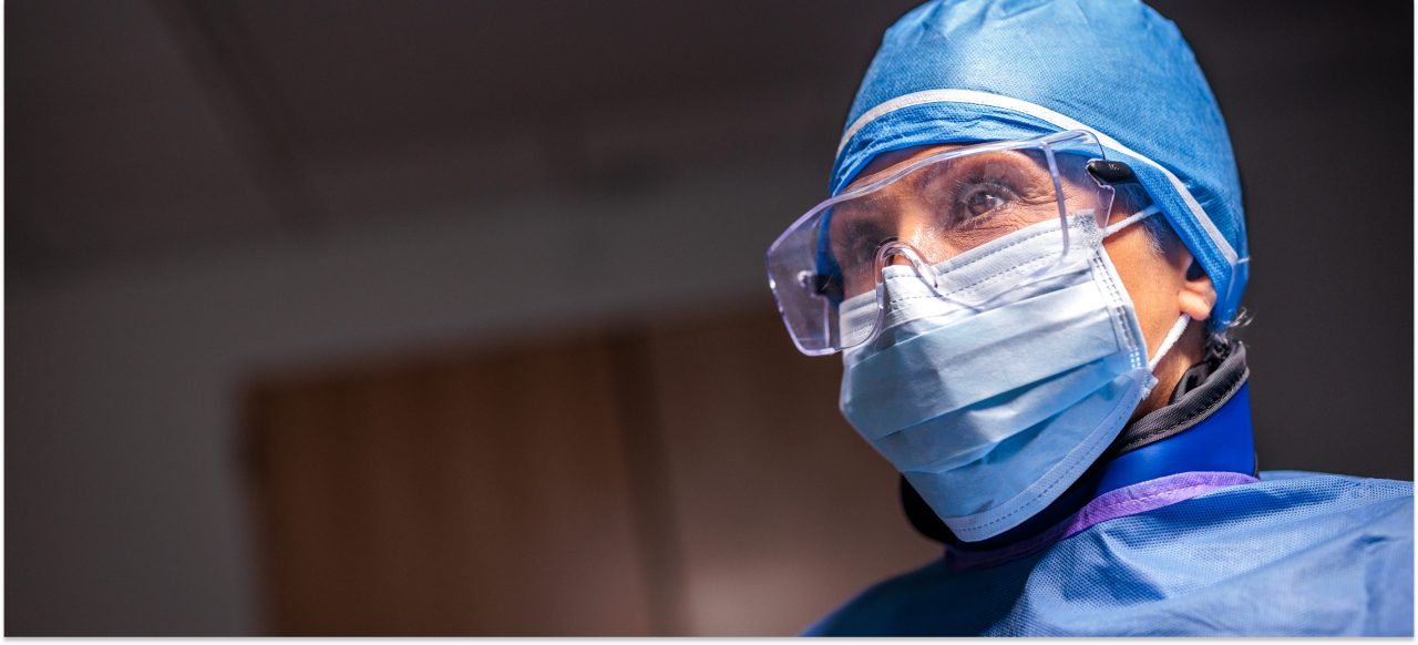 An image of a healthcare prfoessional in blue PPE including cap, eye protection, mask, and gown.