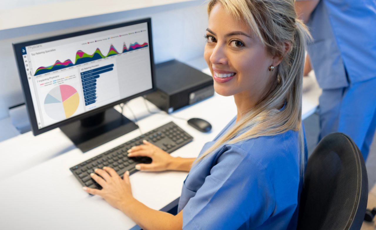 A smiling healthcare professional working at a computer with charts and data on the screen.
