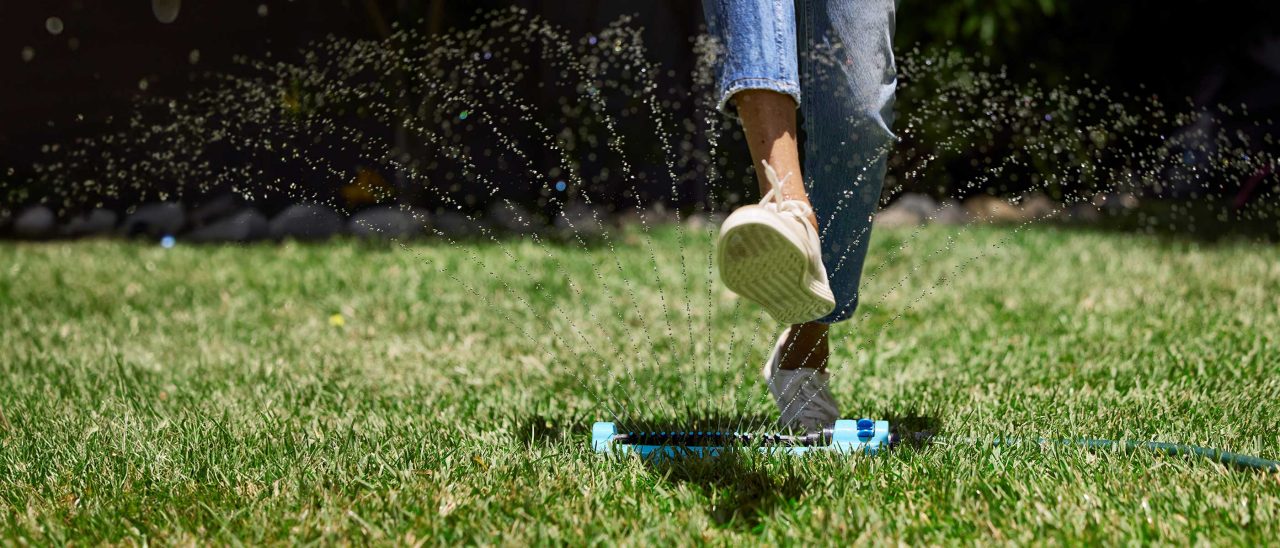 Legs stepping over a running lawn sprinkler.