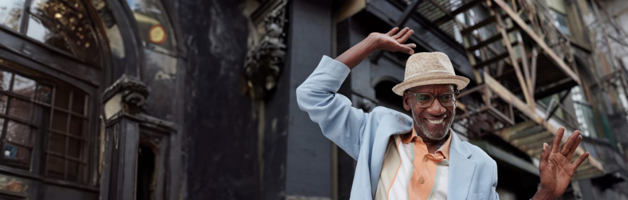 A smiling man dancing in the street wearing a hat.