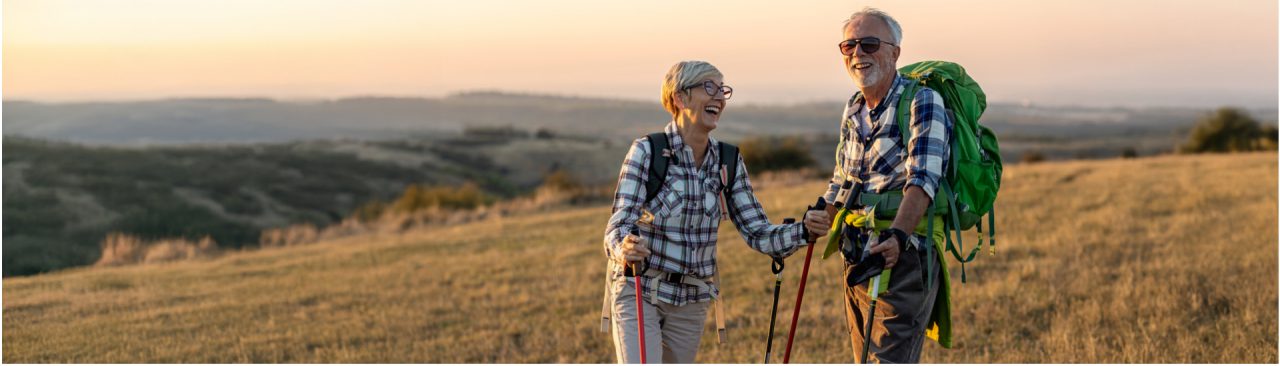 Two smiling people on a hike standing in a grassy field during a sunset.